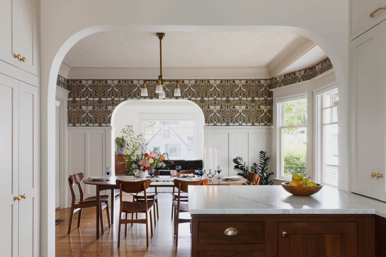 Kitchen with dark stained cabinets looked through arched doorway to dining room with wallpaper.